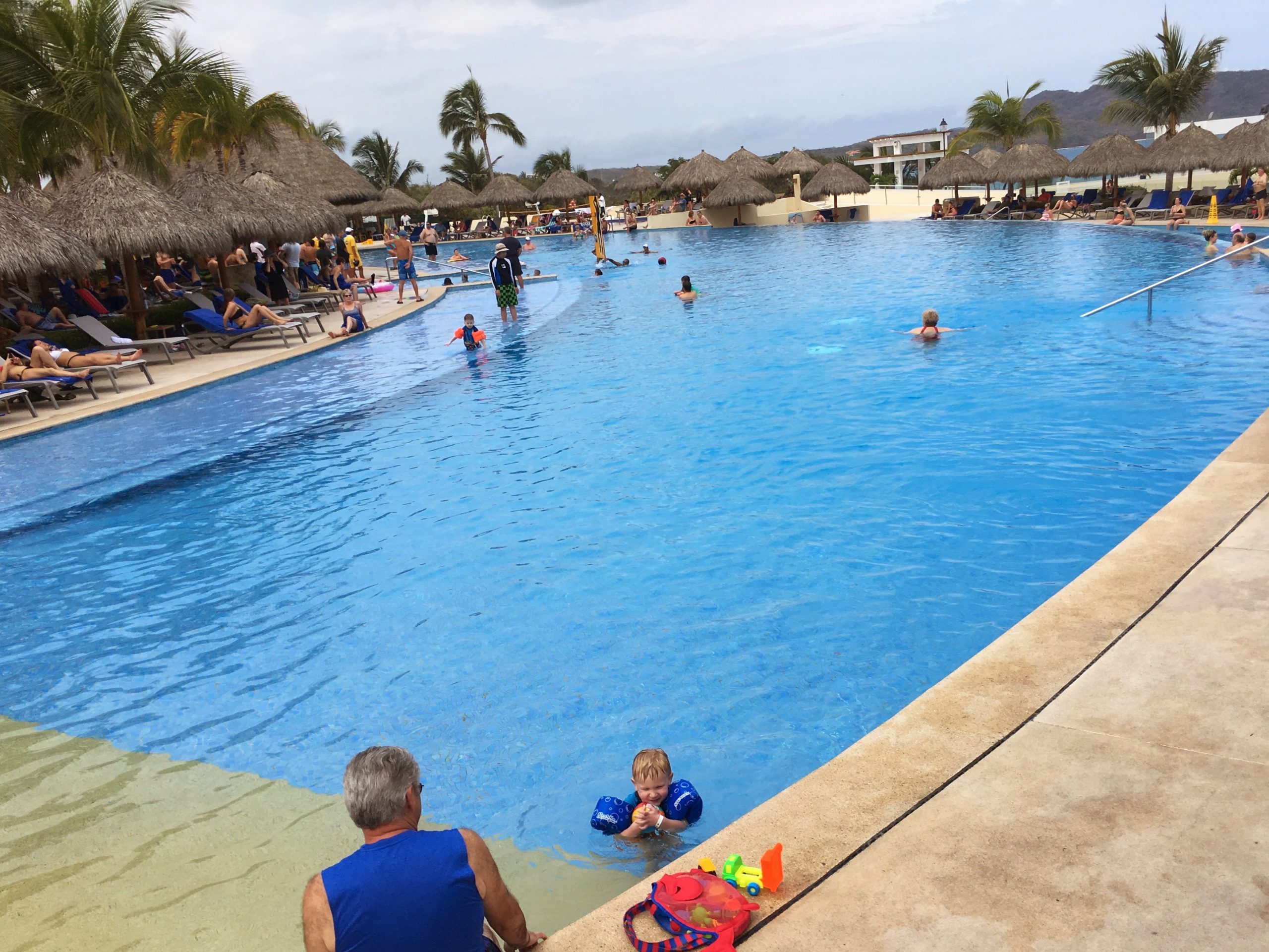 main swimming pool area with beach volleyball set up and a little boy playing in the water